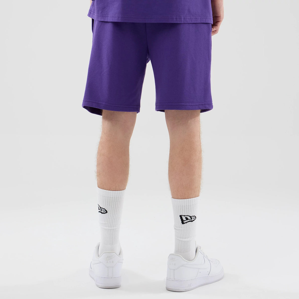 New Era LA Lakers NBA League Essential Purple Oversized Shorts, back view on a model styled with a matching purple top, showing a hemline and relaxed fit ending just above the knee, paired with white socks featuring an embroidered New Era logo.