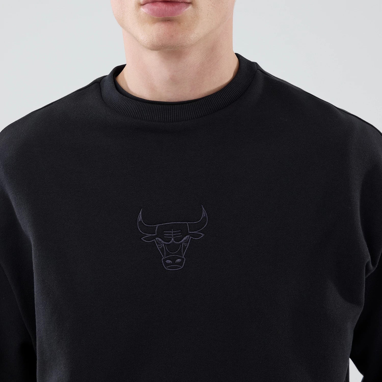 New Era Chicago Bulls NBA League Essential Black Oversized Crew Neck Sweater, front view on a model showing the embroidered black Bulls logo centred on the chest, with a round ribbed collar and loose fit shoulders.