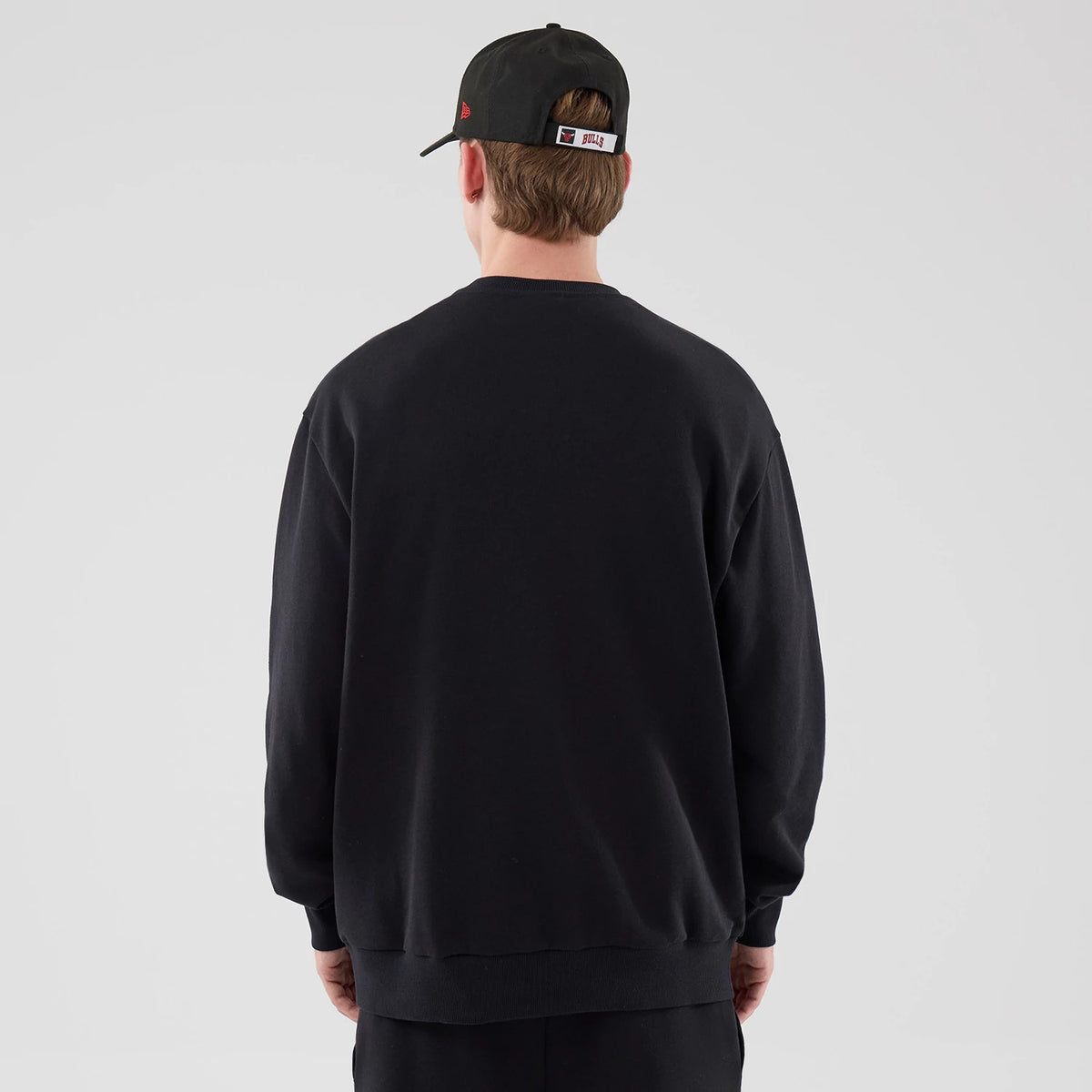 New Era Chicago Bulls NBA League Essential Black Oversized Crew Neck Sweater, back view on a model wearing a black cap, showing the plain black fabric with relaxed shoulders and a loose fit, extending slightly below the waist.