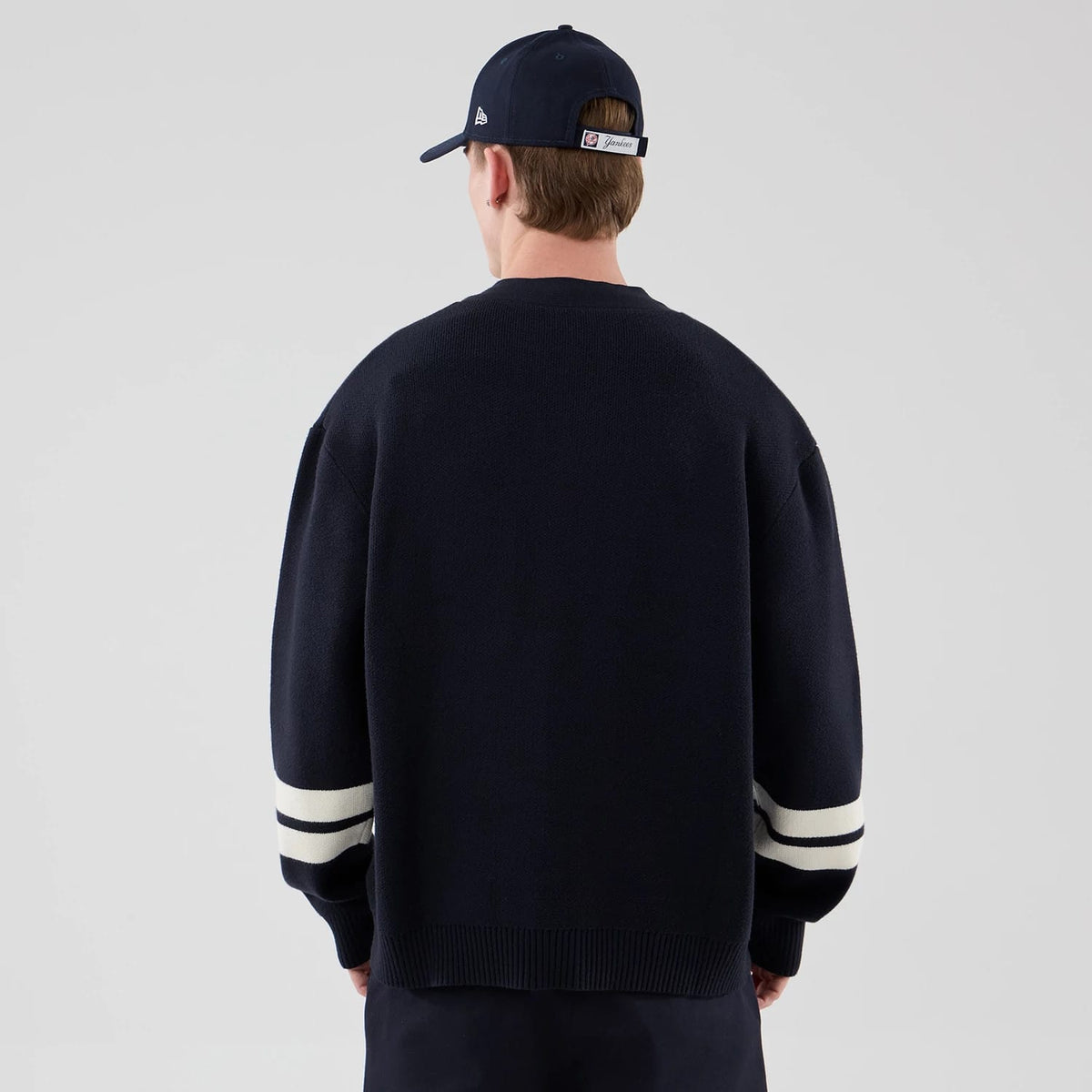 New Era New York Yankees MLB Knit Navy Cardigan, back view on a model also wearing a cap, showing the ribbed hem and cuffs, with two white stripes on each sleeve contrasting against the deep navy knitted fabric.