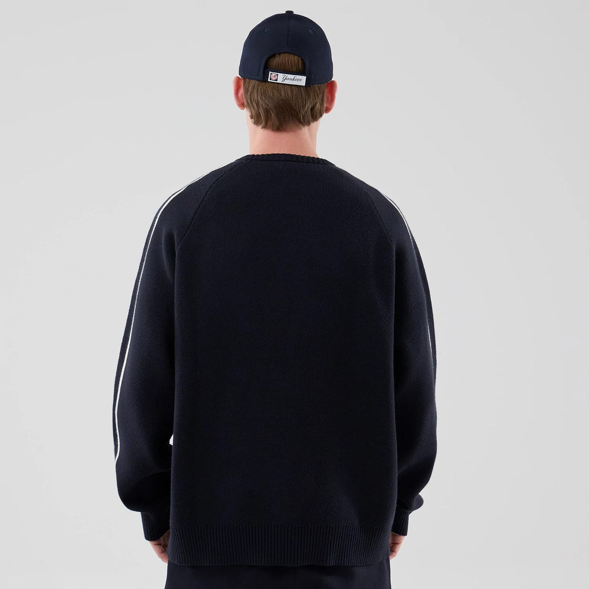 New Era New York Yankees MLB Knit Navy Knitted Sweater, back view on a model wearing a navy cap, with distinct white stripes running along the sleeves, a ribbed hemline, and a soft textured fabric displaying a classic baseball-inspired design.
