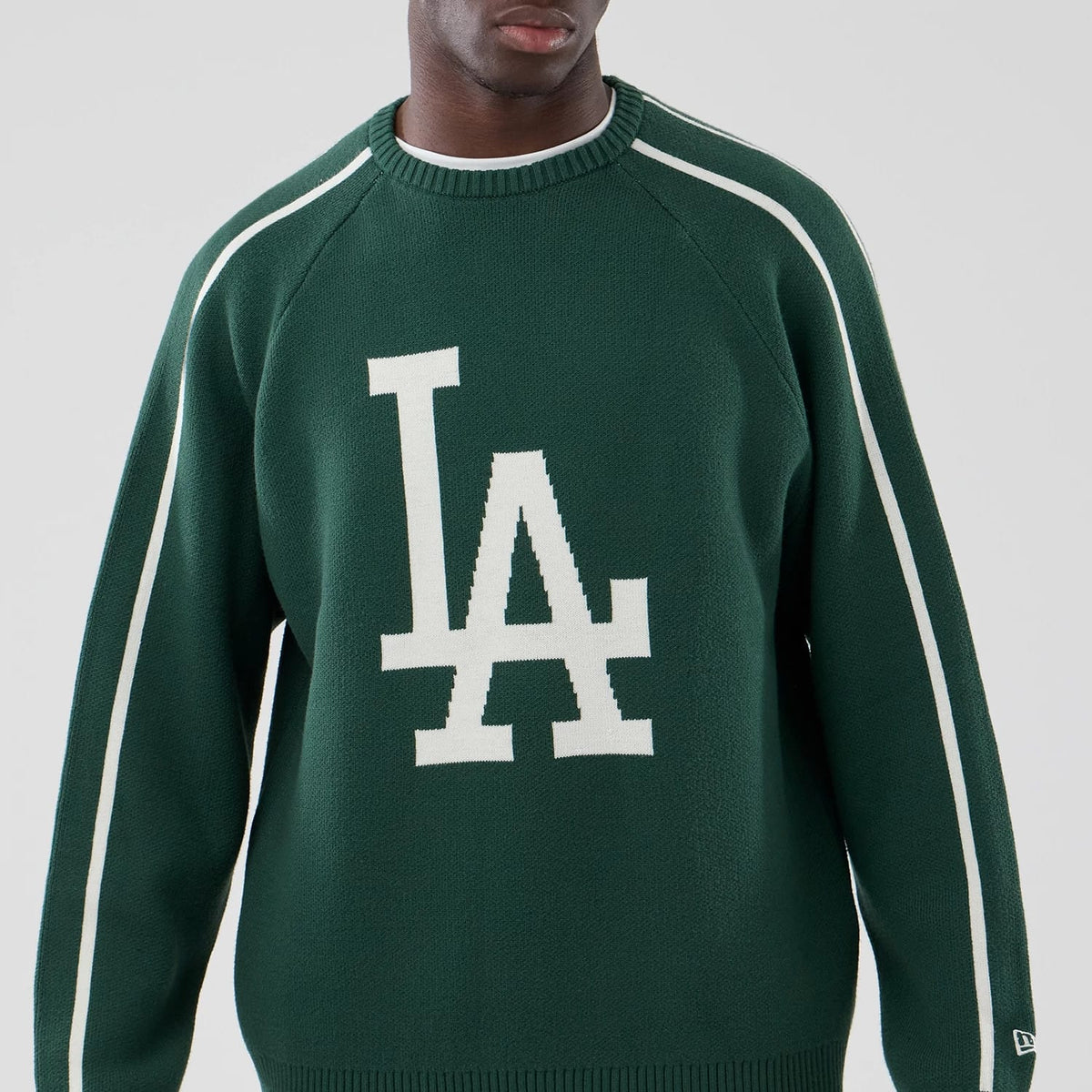 New Era LA Dodgers MLB Knit Dark Green Knitted Sweater, front view on a model showcasing the large white LA logo at the centre of the chest, with contrasting white stripes running down the arms and a ribbed crew neck.