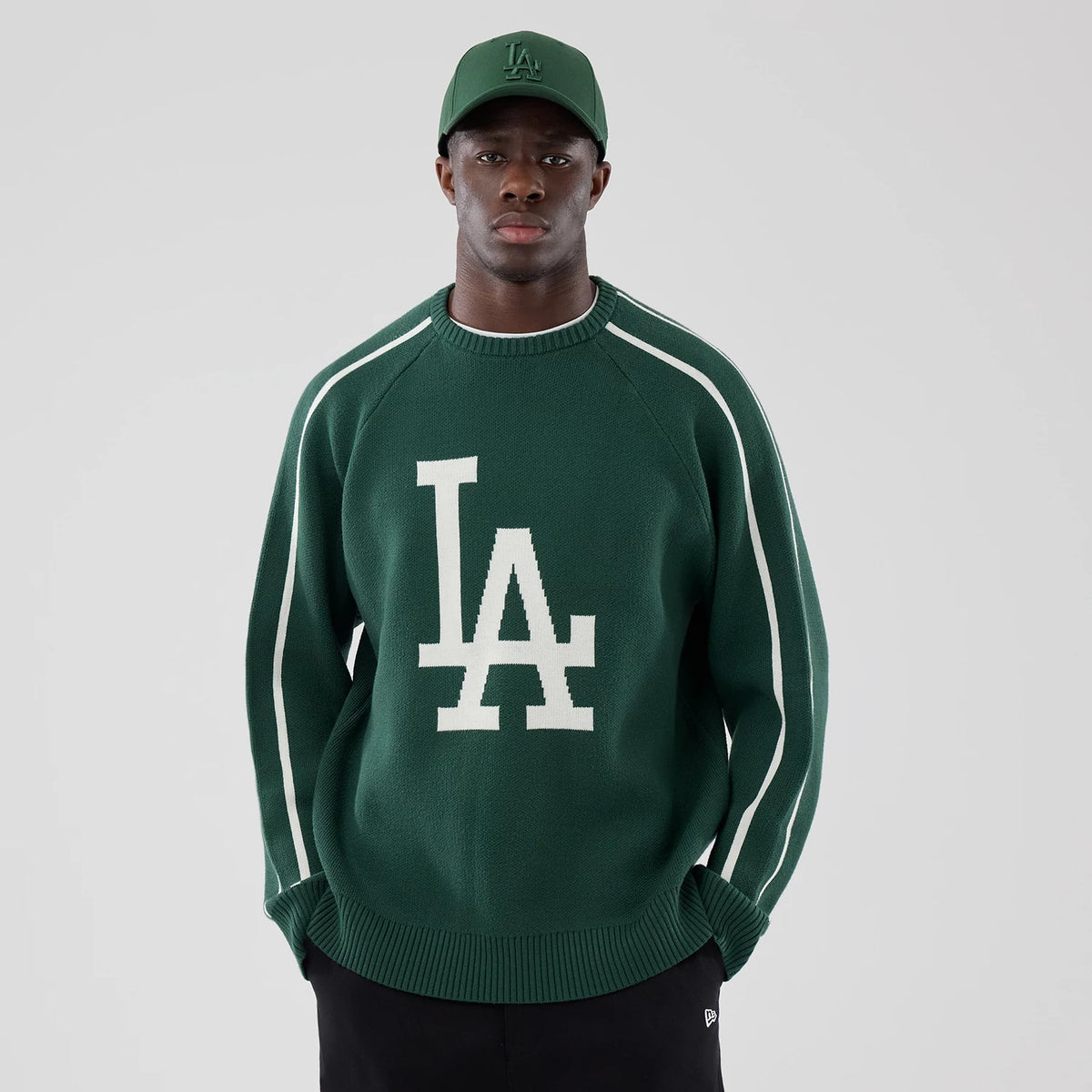 New Era LA Dodgers MLB Knit Dark Green Knitted Sweater, on-model front view showing a large white LA logo knitted on the chest, contrast white stripes on the sleeves, and paired with a matching dark green cap featuring the same logo.