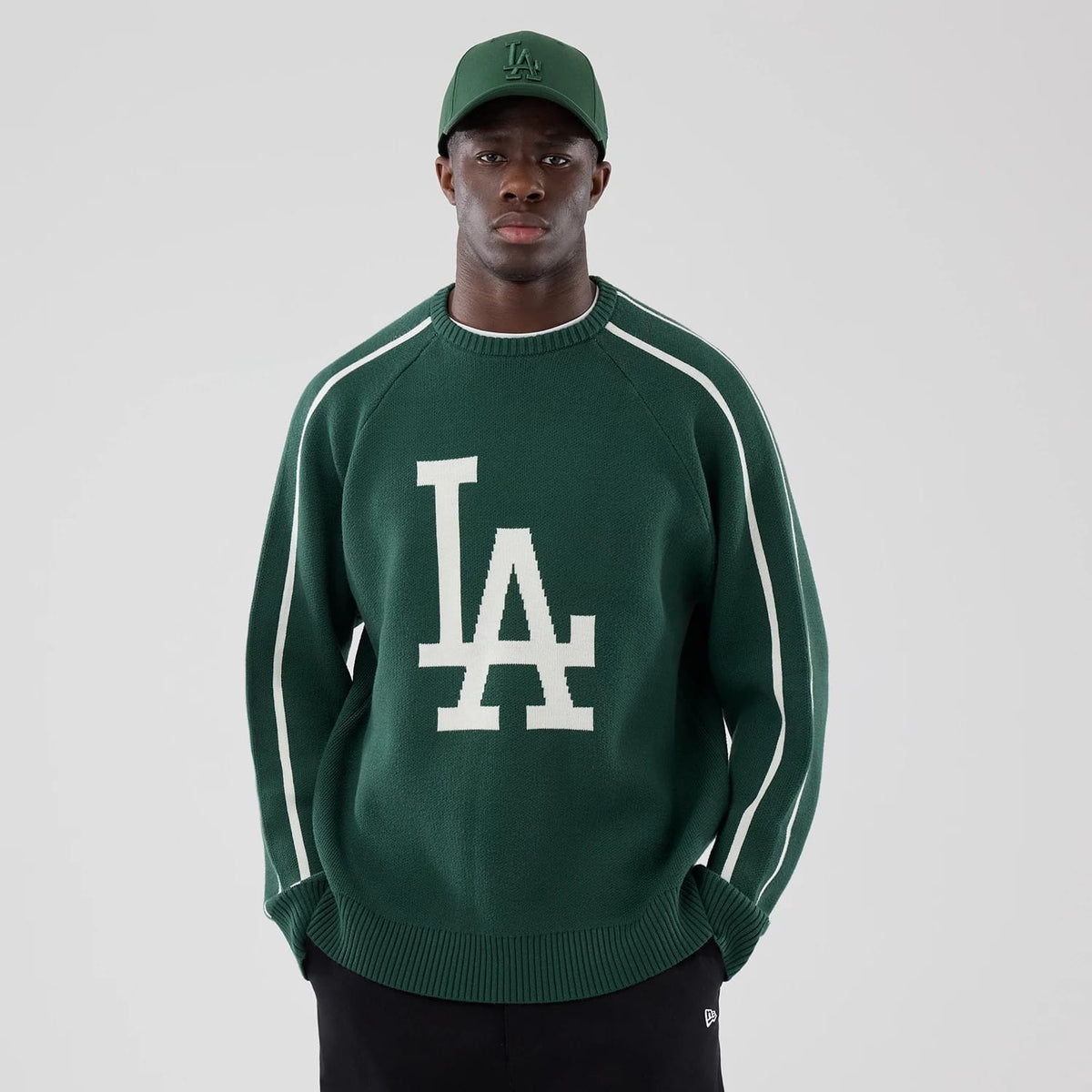 New Era LA Dodgers MLB Knit Dark Green Knitted Sweater, on-model front view showing a large white LA logo knitted on the chest, contrast white stripes on the sleeves, and paired with a matching dark green cap featuring the same logo.