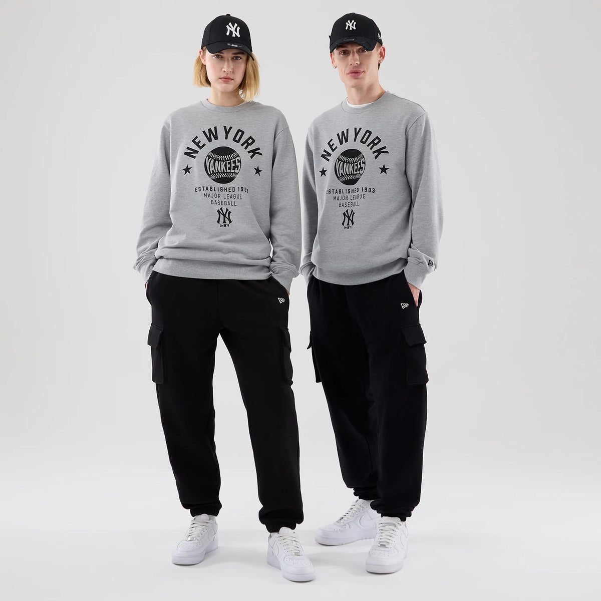New Era New York Yankees MLB Graphic Grey Oversized Crew Neck Sweater, front view on a model wearing a black New York Yankees cap, showcasing black New York Yankees graphics on the chest, paired with black cargo trousers and white trainers.