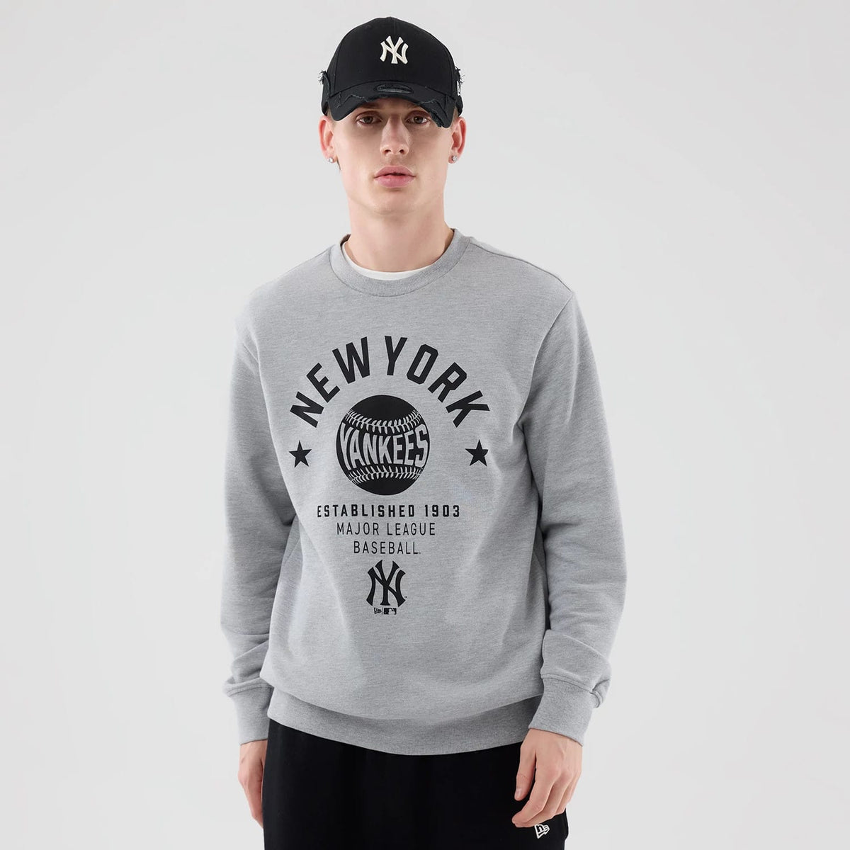 New Era New York Yankees MLB Graphic Grey Oversized Crew Neck Sweater, front view on a model with a New York Yankees cap, featuring prominent black New York Yankees lettering and logo across the chest, with ribbed cuffs and hem.