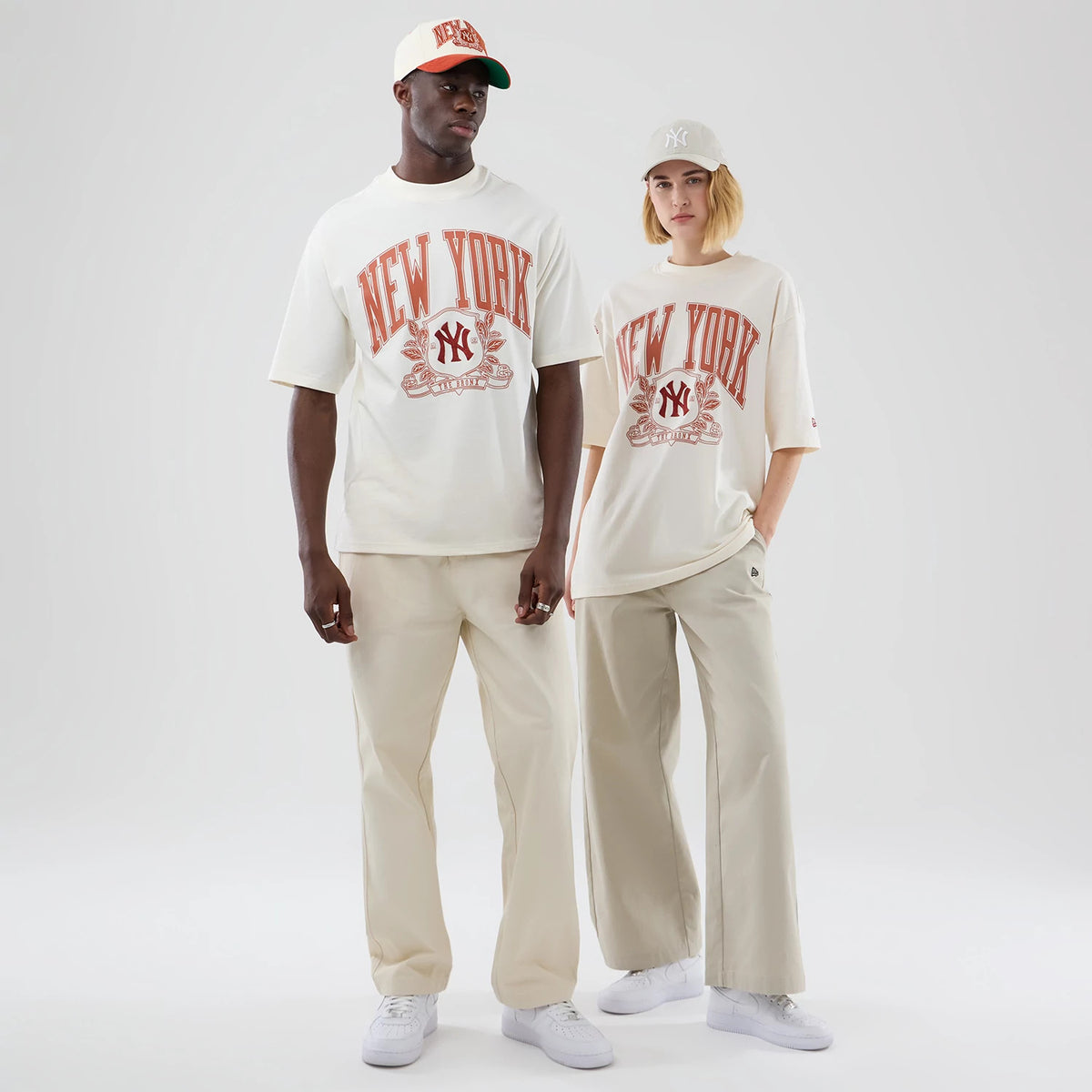 New Era New York Yankees MLB Arch Graphic Open White Oversized T-Shirt, front view on models, displaying the red NEW YORK lettering and Yankees logo on the centre chest, styled with light-coloured trousers and white trainers for a casual look.