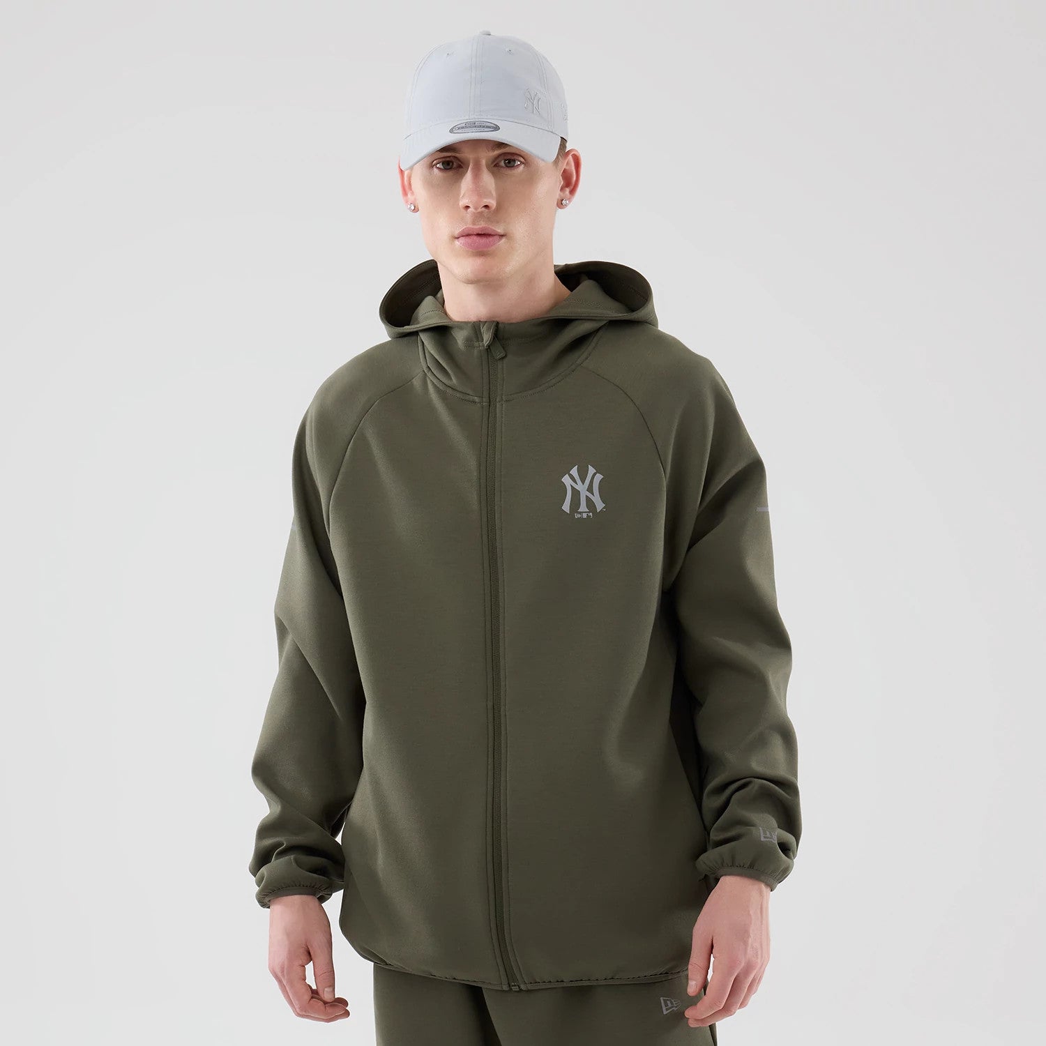 New Era New York Yankees MLB Core Plus Green Full Zip Hoodie, front view on-model showing the olive green hoodie with a grey embroidered New York Yankees logo on the chest, a full-length zip closure, and relaxed fit styled with a light grey cap.