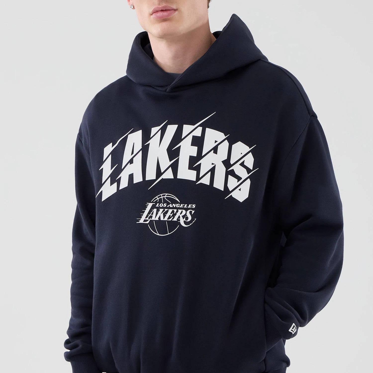 New Era LA Lakers NBA Core Plus Navy Pullover Hoodie, front view on model showcasing a white Lakers graphic printed across the chest, additional Los Angeles Lakers logo below, and a small embroidered New Era logo on the left sleeve cuff.