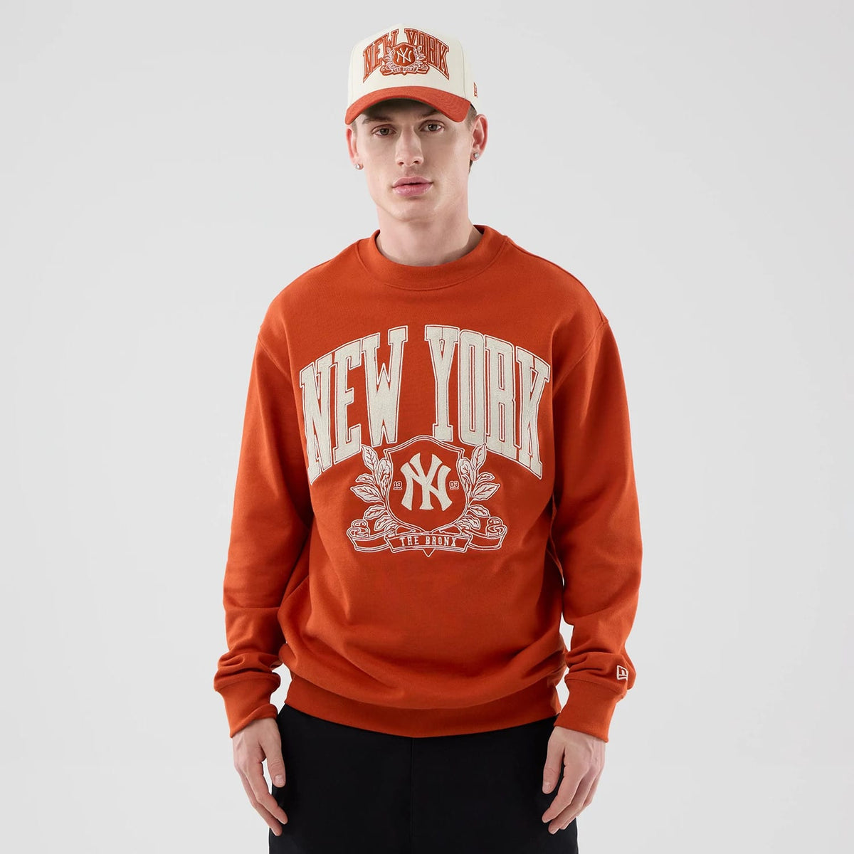 New Era New York Yankees MLB Arch Graphic Orange Oversized Crew Neck Sweater, front view on a model with a matching cap, featuring large New York lettering and the NY logo in white across the chest, along with detailed graphic embroidery.