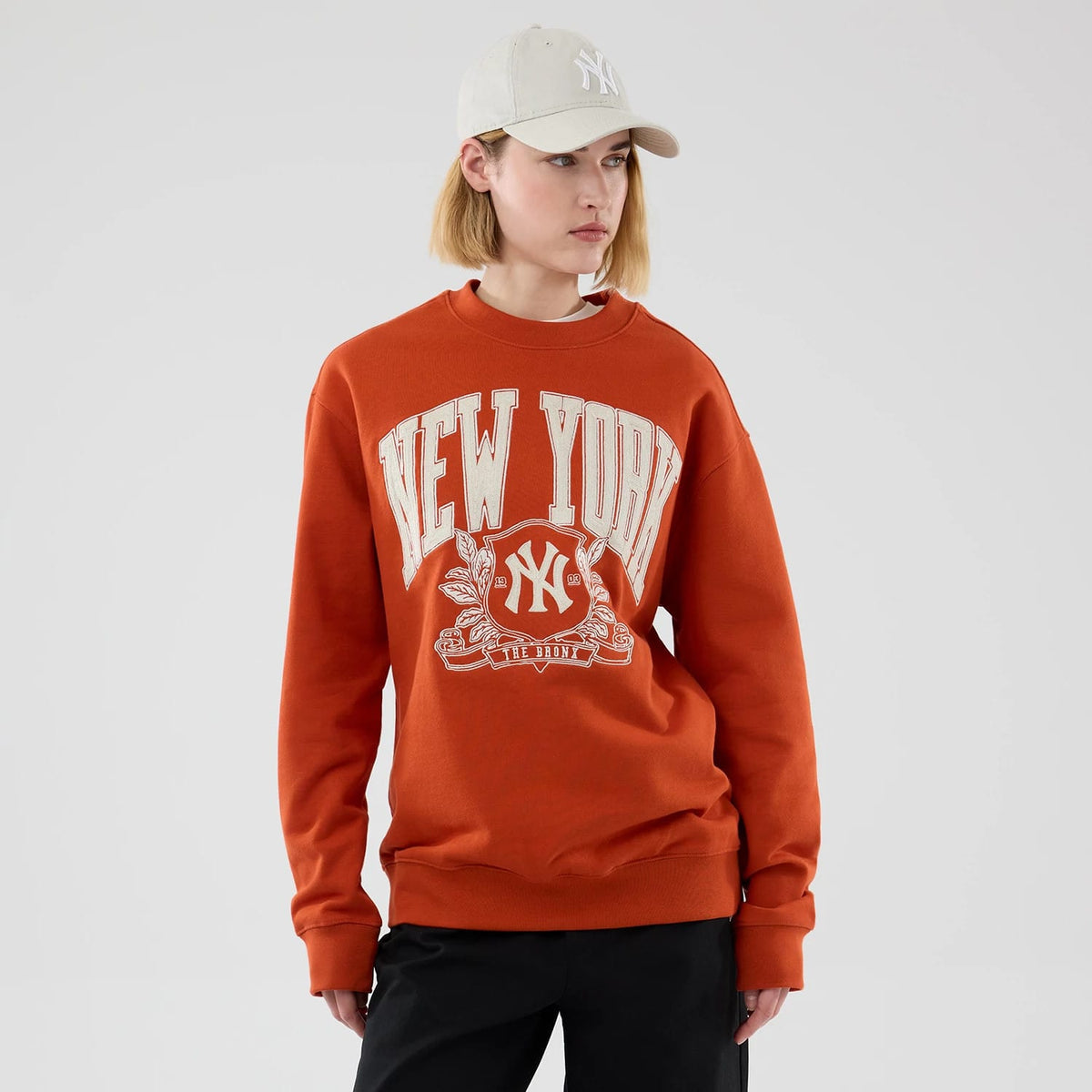New Era New York Yankees MLB Arch Graphic Orange Oversized Crew Neck Sweater, front view on a model showing the large cream 'New York' lettering across the chest, an ornate Yankees NY logo with laurel details, and relaxed fit with ribbed cuffs.