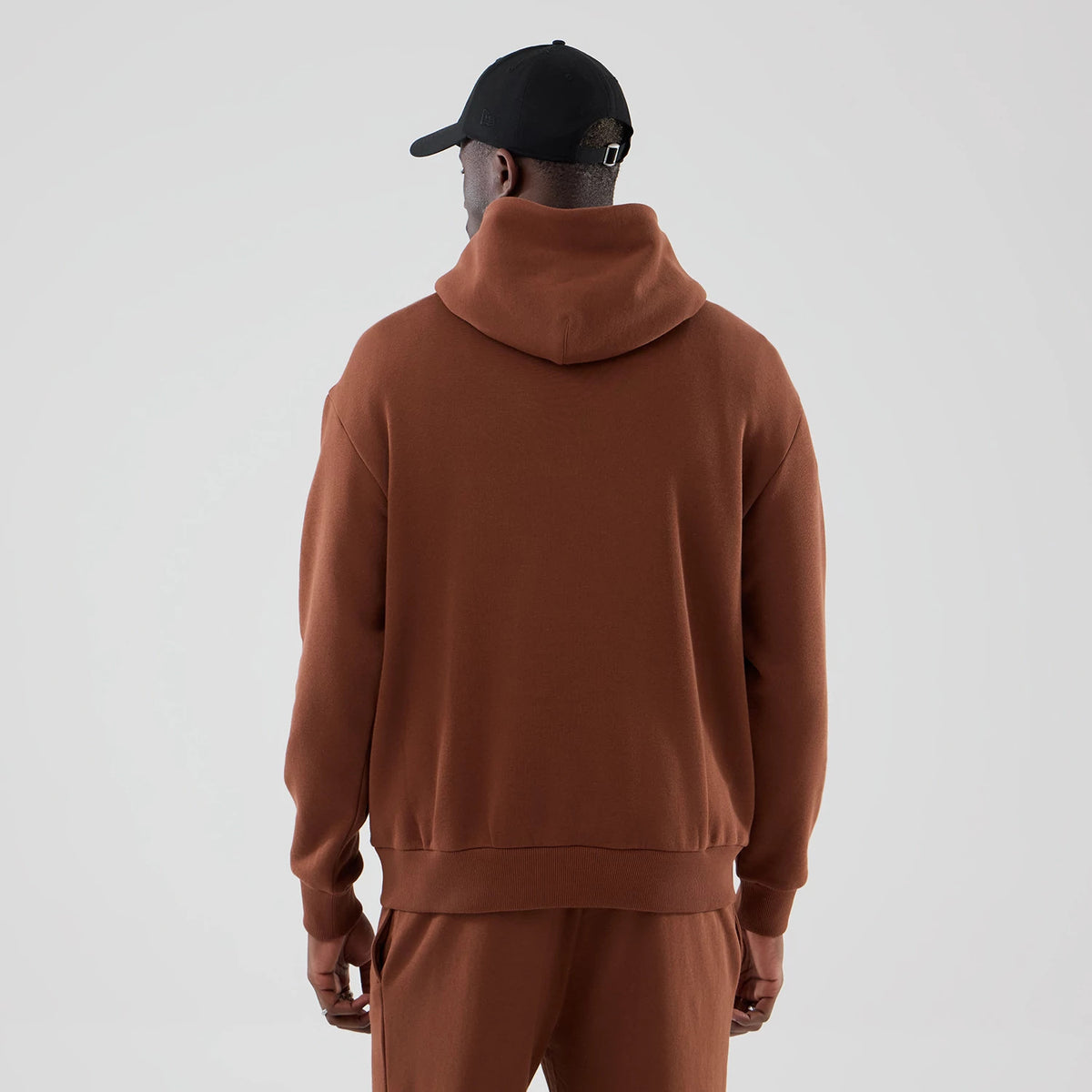 New Era Chicago Bulls NBA Core Plus Brown Pullover Hoodie, back view on a model wearing matching brown trousers and a black cap, showing a plain brown design with a spacious hood and ribbed cuffs, hem sitting at the waistline.