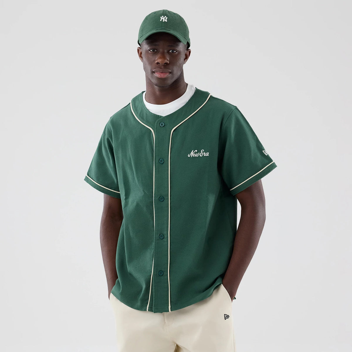 New Era Dark Green Baseball Jersey, front view on a model showing the New Era script logo embroidered in white on the left chest, contrasting white piping along the button placket and cuffs, and a matching dark green fabric with short sleeves.