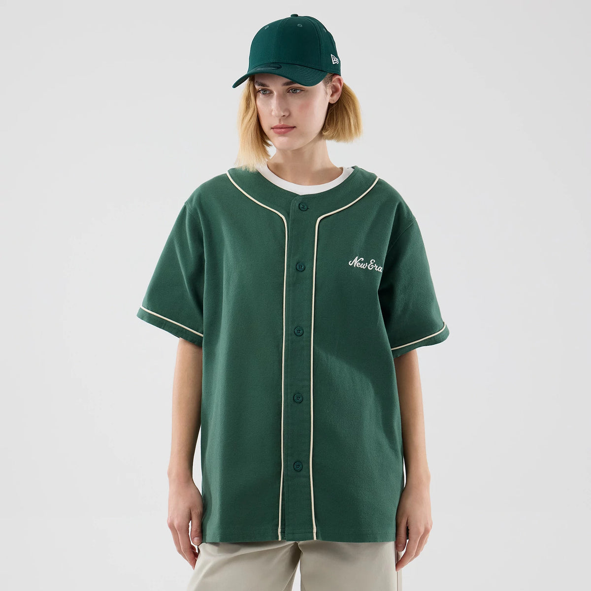 New Era Dark Green Baseball Jersey, front view on a model with a green cap, featuring cream piping along the button placket and sleeve edges, an embroidered New Era script logo on the left chest, and a relaxed fit.