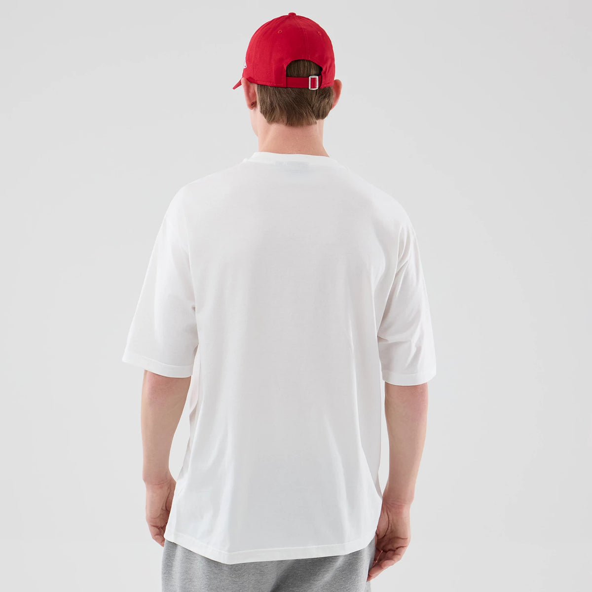 New Era Chicago Bulls NBA Script Graphic White Oversized T-Shirt, back view on a model wearing a red cap and grey shorts, displaying a plain white back with loose-fit short sleeves and slightly dropped shoulders.