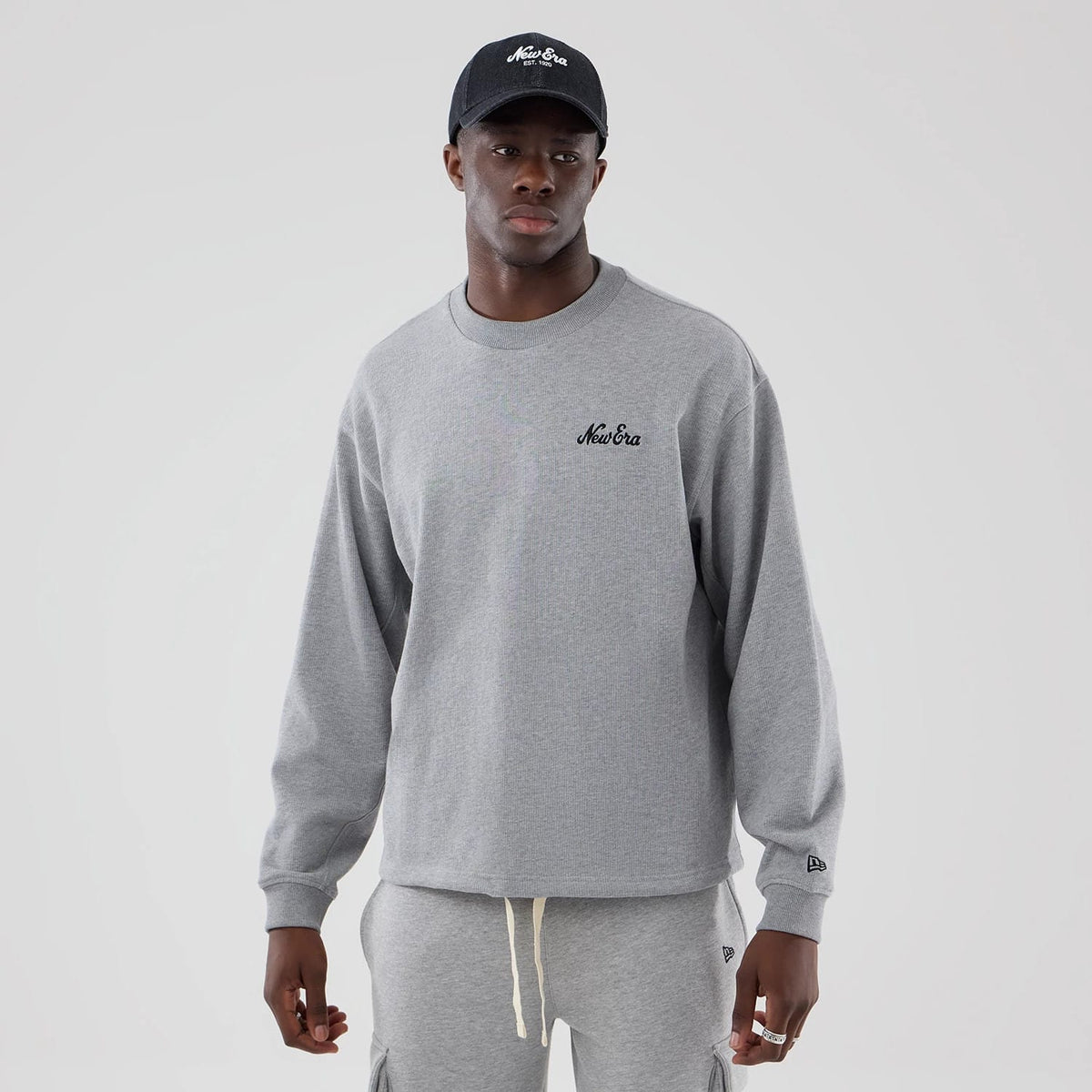 New Era Grey Oversized Crew Neck Sweater, front view on a model with a black cap, featuring the New Era script logo embroidered in black on the left chest, relaxed fit sleeves, and the New Era flag logo on the left cuff.