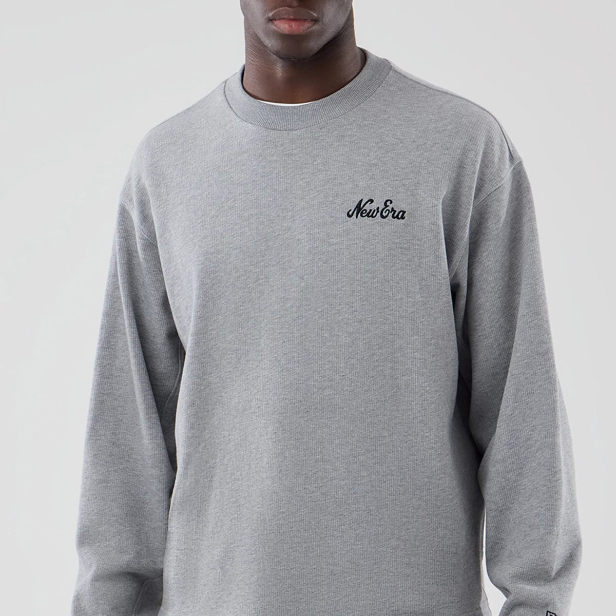 New Era Grey Oversized Crew Neck Sweater, front view on a model showing the black embroidered New Era script logo on the left chest, ribbed knit collar, and long sleeves with a relaxed fit across the shoulders and torso.