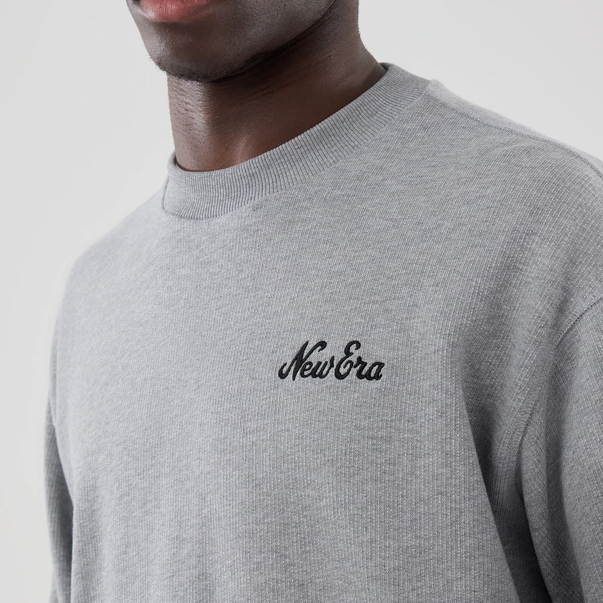 New Era Grey Oversized Crew Neck Sweater, on a model with shoulder and neck, showing the embroidered black New Era logo on the left chest, ribbed crew neck collar, and soft grey fabric texture.