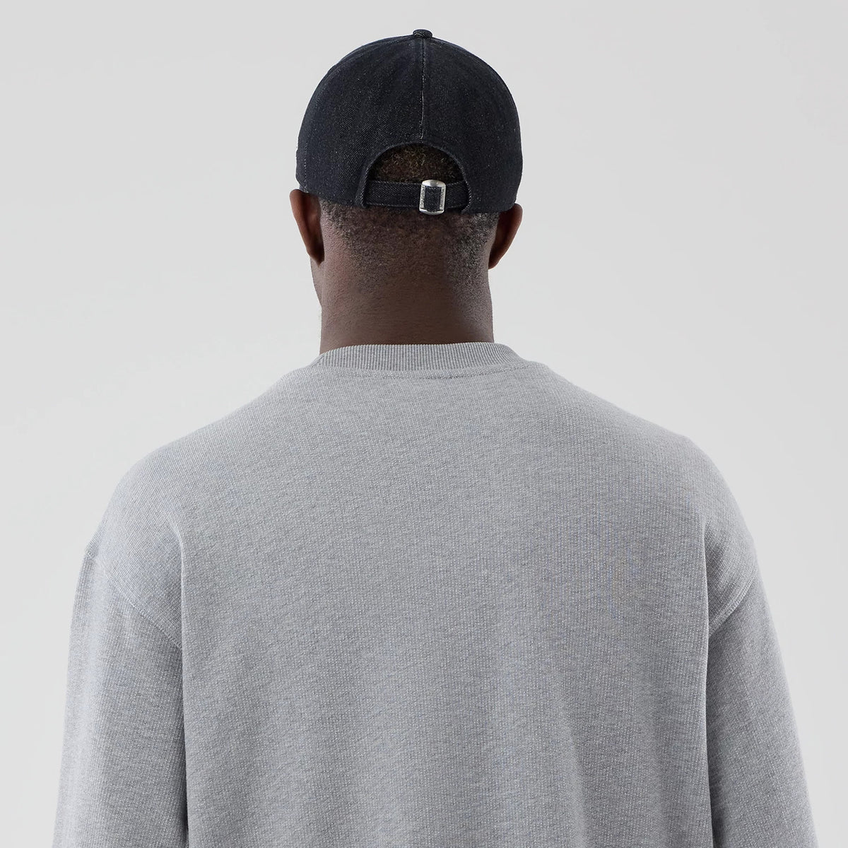 New Era Grey Oversized Crew Neck Sweater, back view on a model wearing a black cap, highlighting the sweater's soft ribbed knit fabric, drop shoulder design, and relaxed fit that drapes comfortably over the shoulders.
