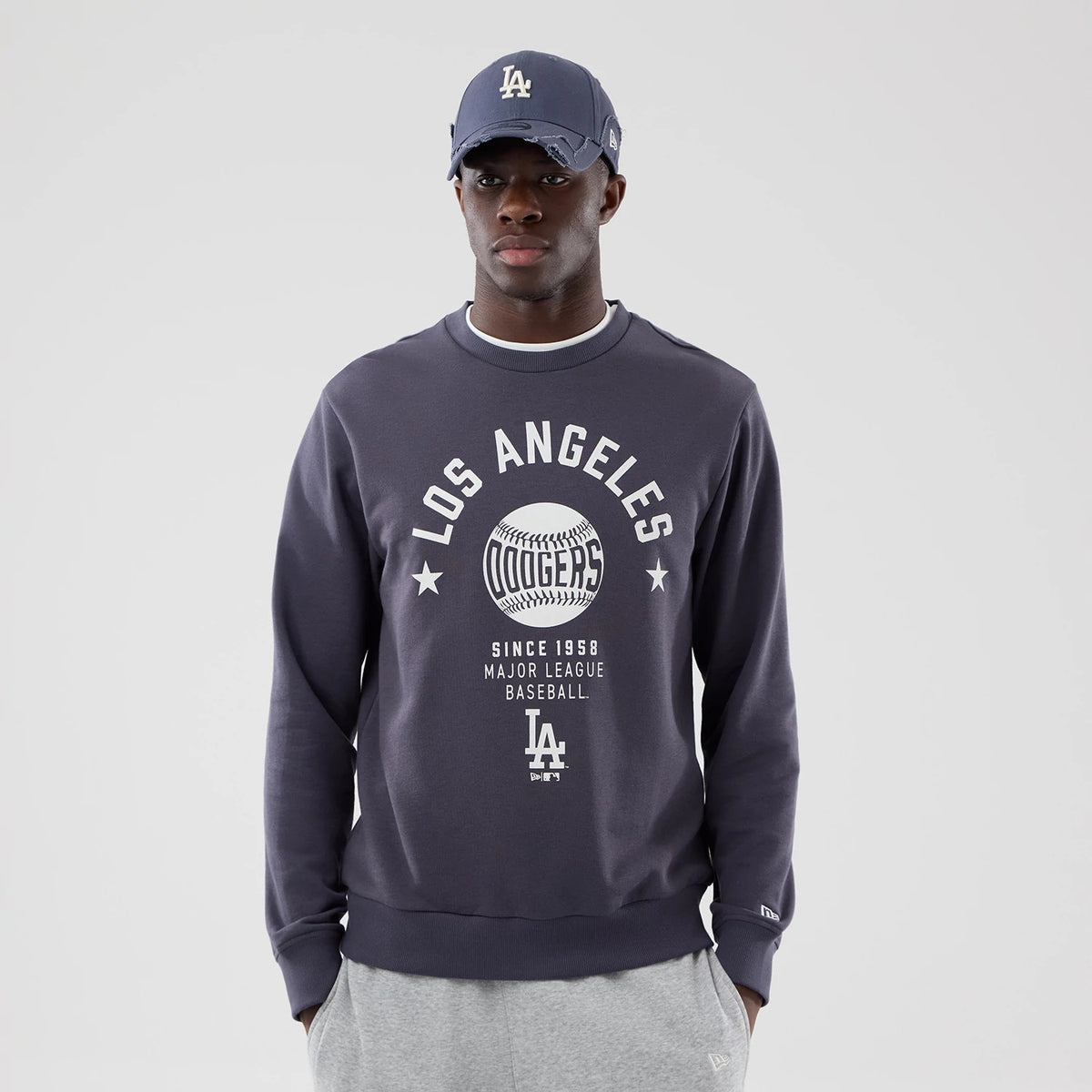New Era LA Dodgers MLB Graphic Grey Oversized Crew Neck Sweater, front view on a model wearing a matching cap, featuring a prominent Los Angeles Dodgers baseball graphic in white across the chest and New Era flag logo on the left sleeve.