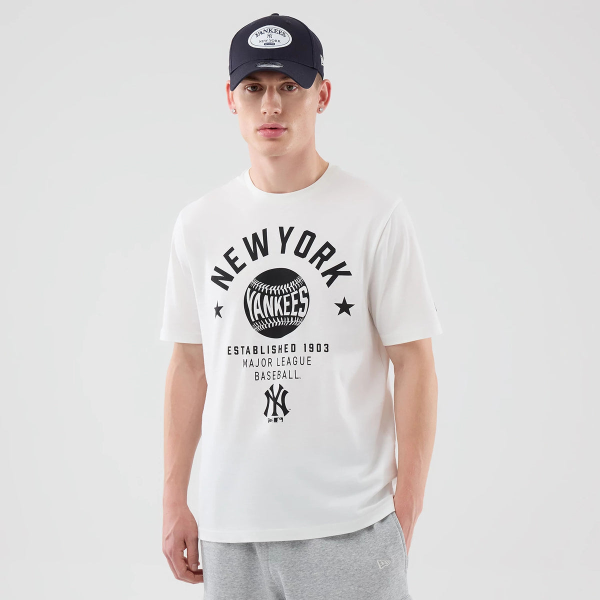 New Era New York Yankees MLB Graphic White T-Shirt, front view on a model with grey trousers and a navy cap, featuring the black printed New York Yankees logo, baseball graphic, and lettering centred on the chest.