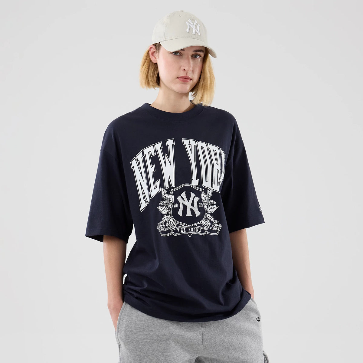 New Era New York Yankees MLB Arch Graphic Navy Oversized T-Shirt, front view on a model styled with grey casual trousers and a matching light cream cap, featuring large white New York lettering across the chest above an embroidered Yankees logo.