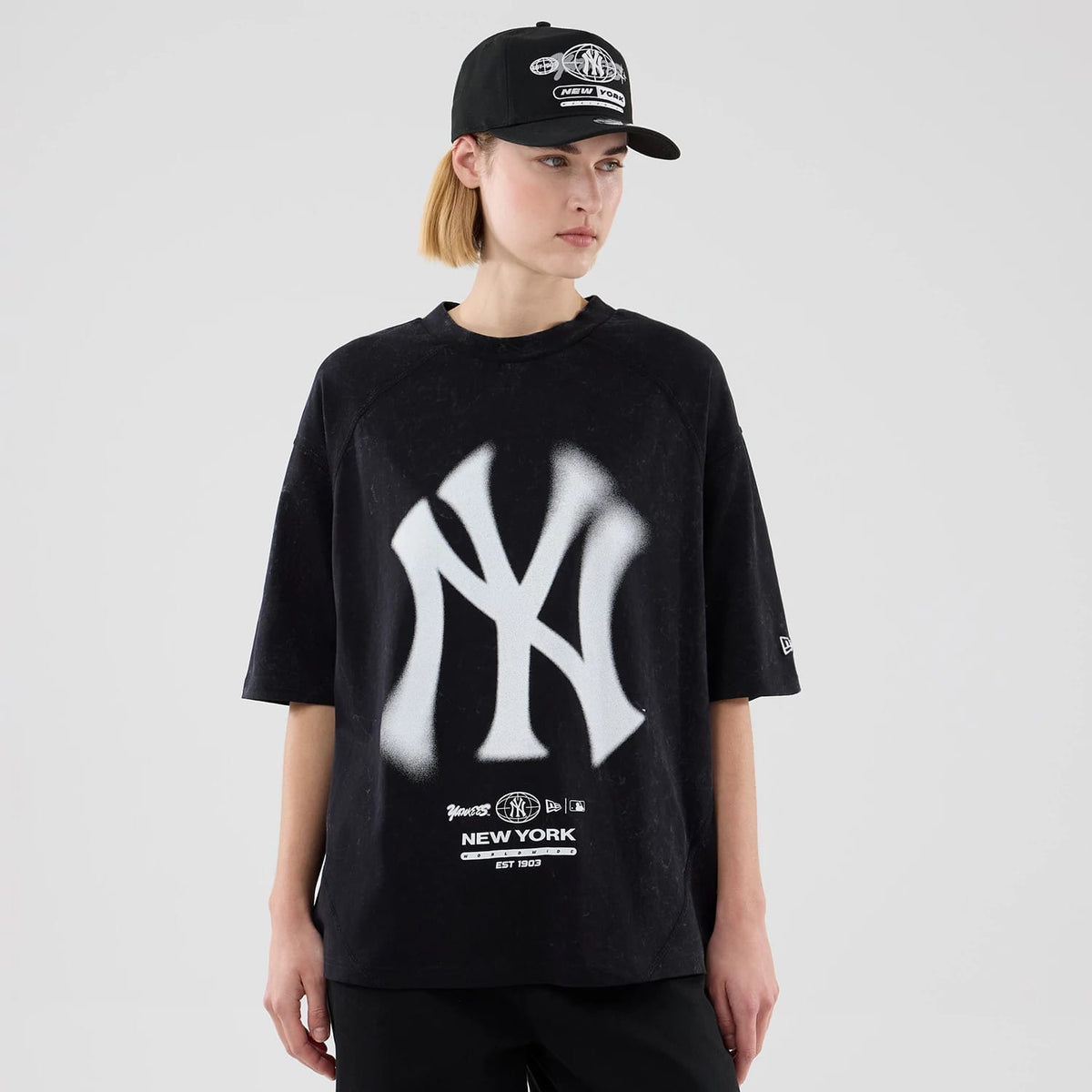 New Era New York Yankees MLB Graphic Boxy Black T-Shirt, front view on a model featuring a large white spray-painted Yankees logo across the chest, smaller team and New Era logos beneath, and an embroidered New Era logo on the left sleeve.