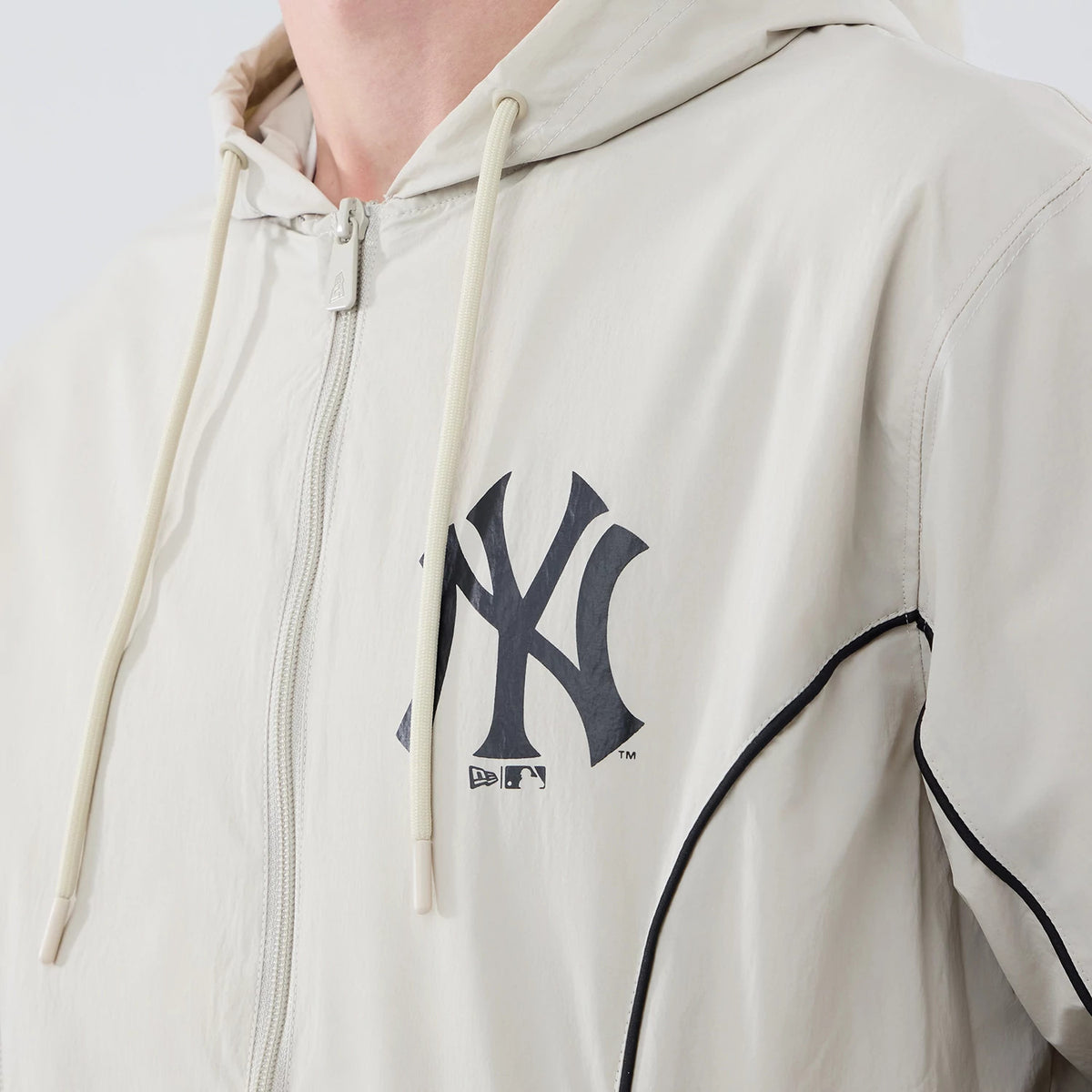 New Era New York Yankees MLB Piping Cream Jacket, three-quarter view showing the large black printed Yankees logo on the left chest, a central zip closure with cream drawstrings at the hood, and distinct black piping running along the shoulder seam.