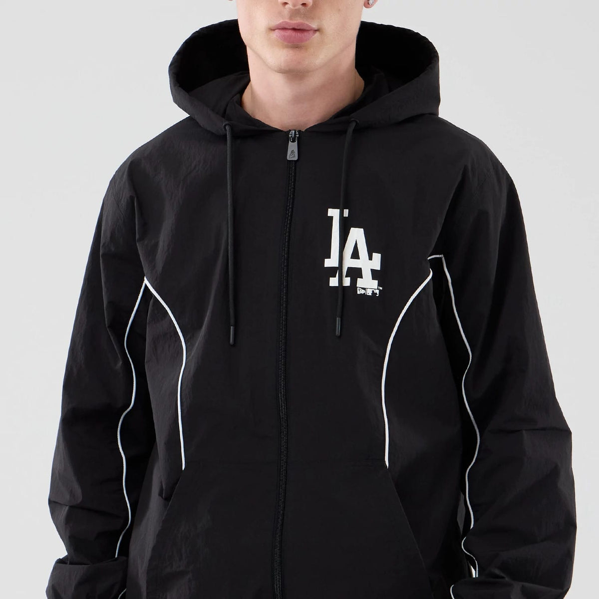 New Era LA Dodgers MLB Piping Black Jacket, front view on a model showing the black jacket with white embroidered LA logo on the left chest, white piping along the sleeves, and a full zip closure with an adjustable drawstring hood.