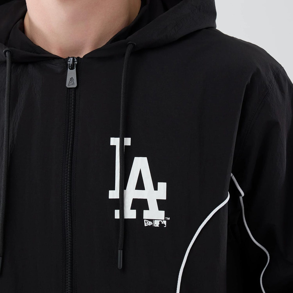New Era LA Dodgers MLB Piping Black Jacket, front view showing the white LA logo printed on the left chest, full-length zip with pull tab at the centre, and distinctive white piping detail running along the shoulders and sides.