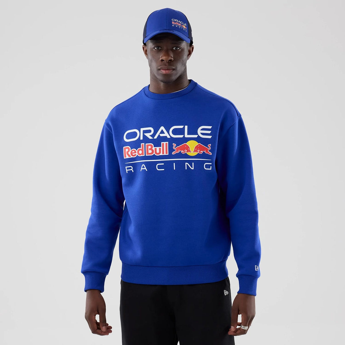 New Era Red Bull Racing Blue Oversized Crew Neck Sweater, front view on a model wearing a matching cap, featuring the Oracle Red Bull Racing logo across the chest in white text with red and yellow details, and ribbed cuffs and hem.