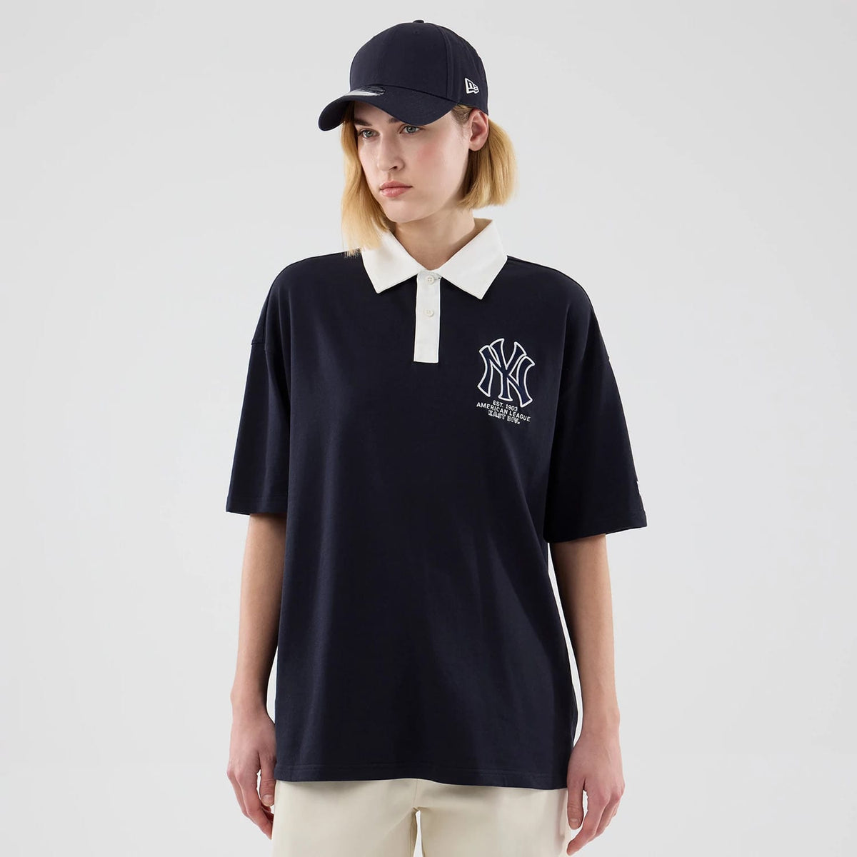 New Era New York Yankees MLB Navy Polo T-Shirt, front view on a model with a matching navy cap, showcasing a white embroidered Yankees logo on the left chest, a white collar with buttons, and short sleeves hanging loosely.