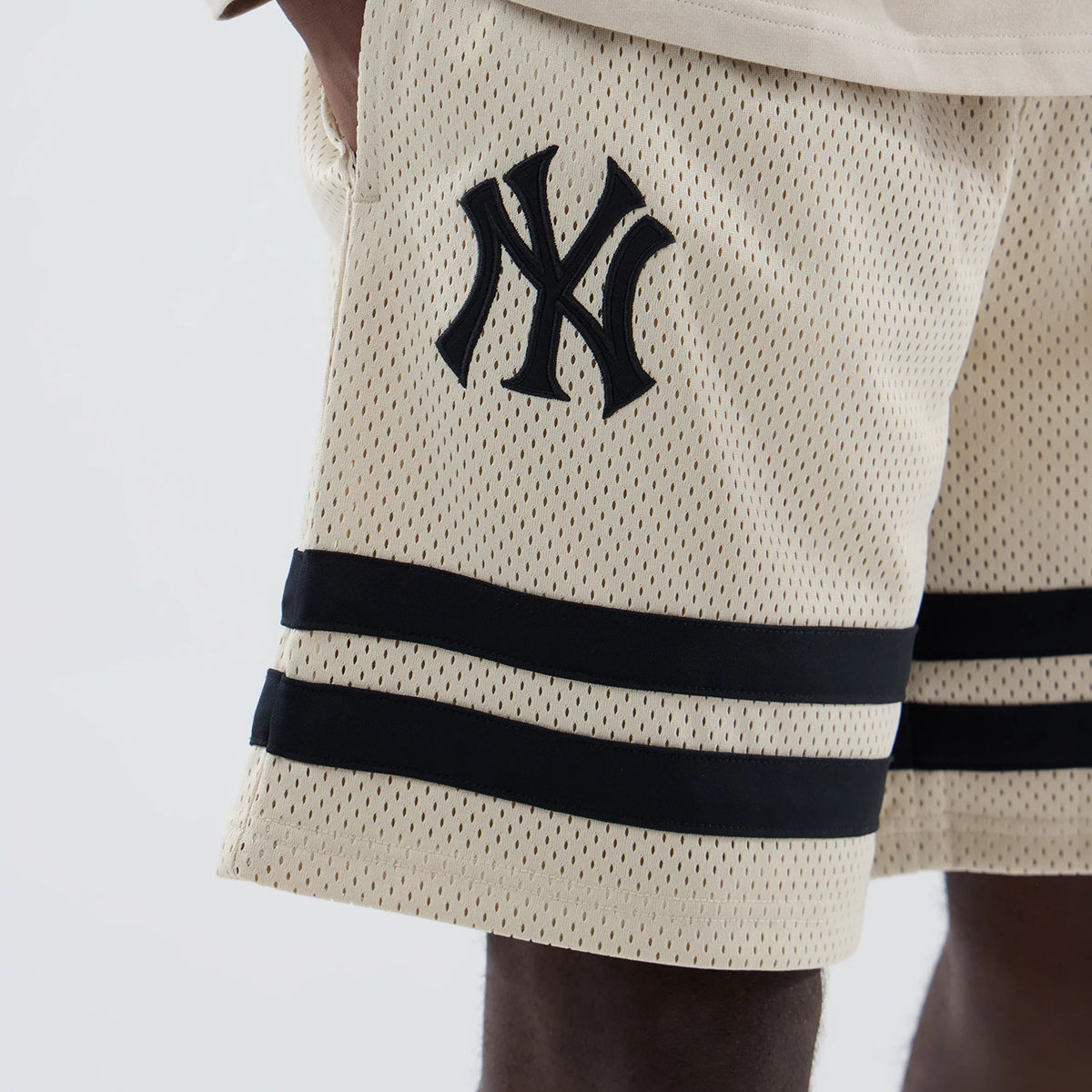 New Era New York Yankees MLB Light Beige Oversized Mesh Shorts, side view showing the embroidered black New York Yankees logo on the upper front, double black horizontal stripes running near the hem, and light beige mesh fabric throughout.