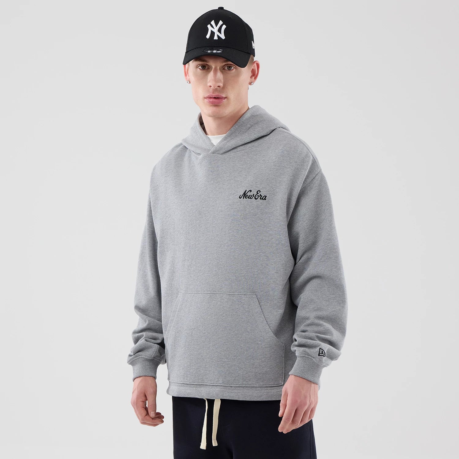 New Era Open Hem Grey Pullover Hoodie, front view on-model showing the embroidered New Era logo on the left chest, a large kangaroo pocket at the front, and branding on the left cuff, styled with a black New York Yankees cap.