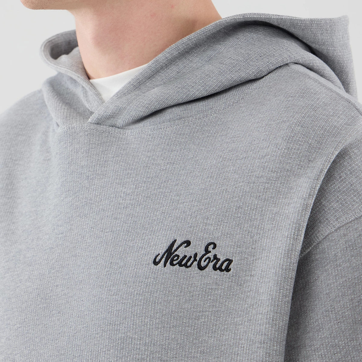 New Era Open Hem Grey Pullover Hoodie, three-quarter view showing the grey fabric, with an embroidered black New Era logo on the left chest, featuring a relaxed hood and worn over a white t-shirt at the neckline.