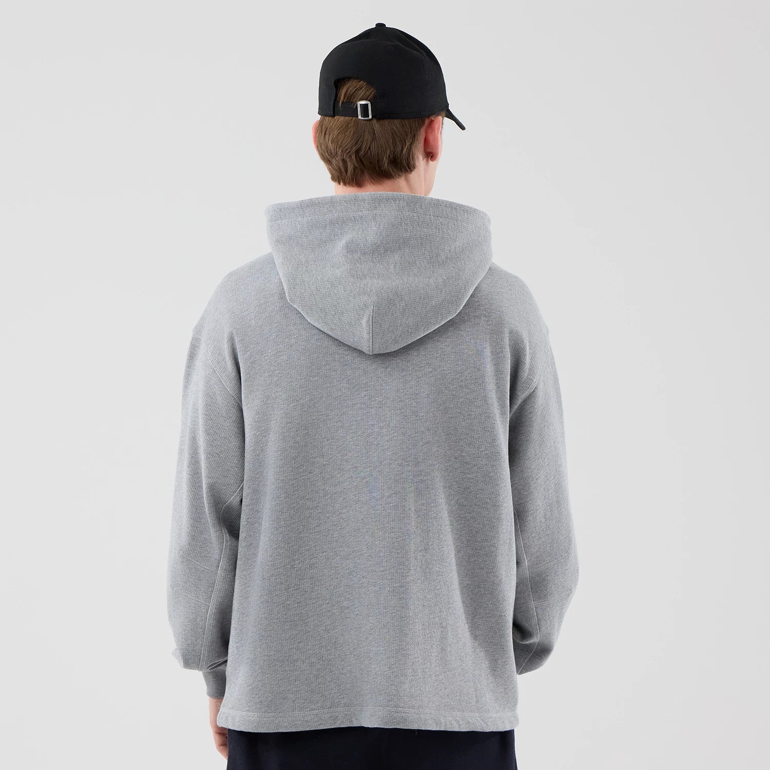 New Era Open Hem Grey Pullover Hoodie, back view on a model wearing a black cap, displaying the relaxed fit and spacious hood, extending over the shoulders, with long sleeves and a clean, uninterrupted grey back design.