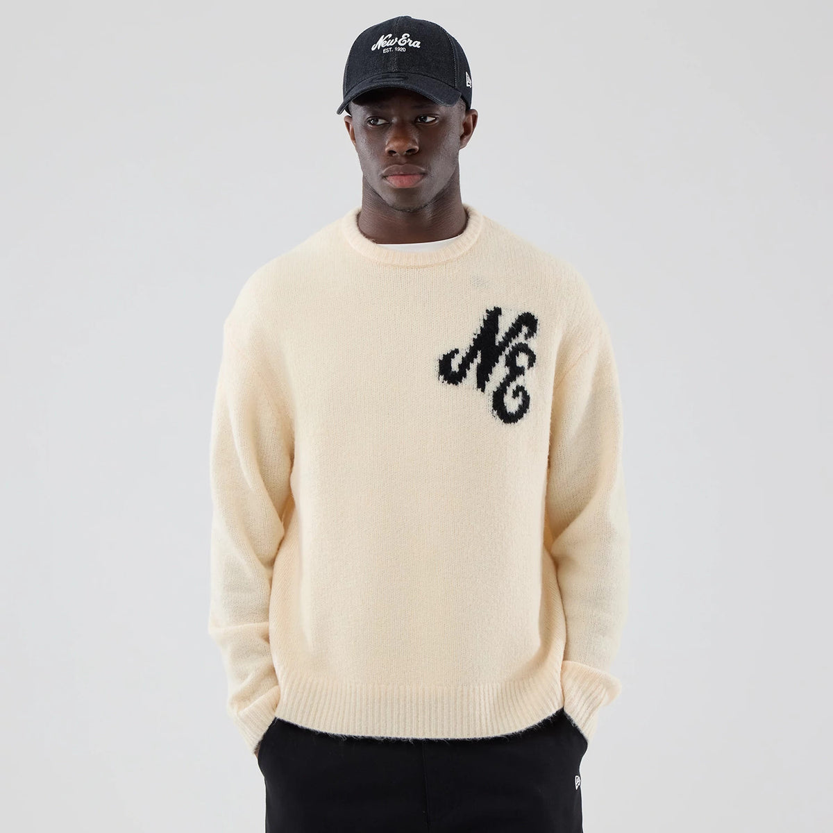 New Era Knit Cream Knitted Sweater, front view on a model wearing a matching black cap, featuring a large black NE knitted logo on the left chest, a ribbed neckline, cuffs, and hem enhancing the soft cream texture.