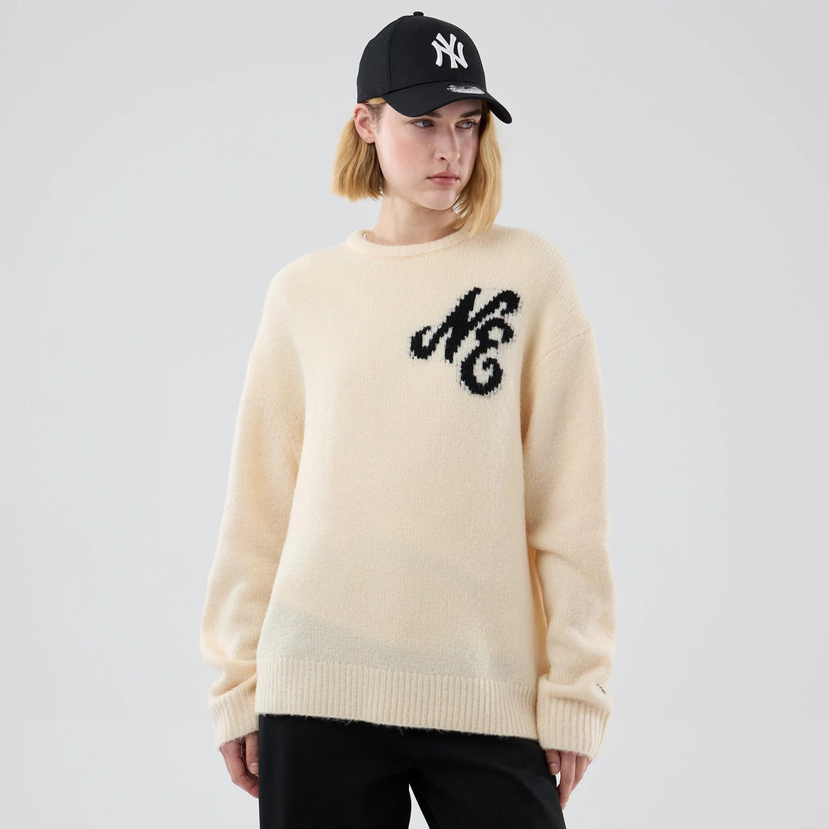 New Era Knit Cream Knitted Sweater, on-model view with a black cap, featuring a black embroidered NE logo on the upper front left, ribbed cuffs and hem, and a soft cream texture that enhances its cosy appearance.