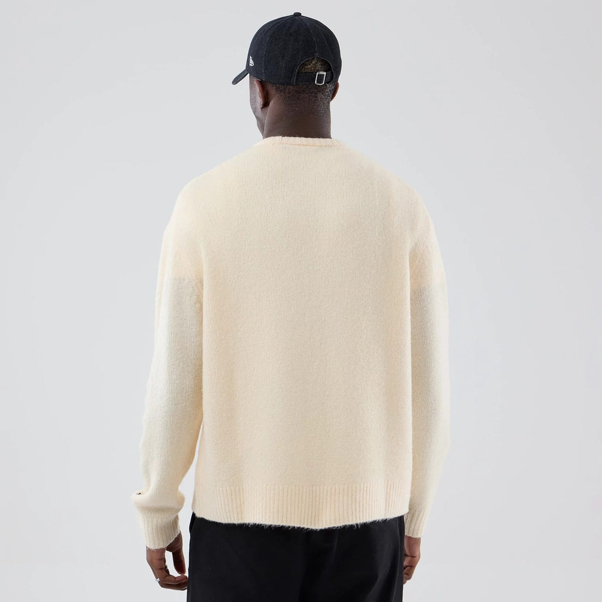 New Era Knit Cream Knitted Sweater, back view on a model wearing a black cap, showing the sweater in a soft cream colour with long sleeves and a ribbed hem detail, highlighting the relaxed fit and textured material.