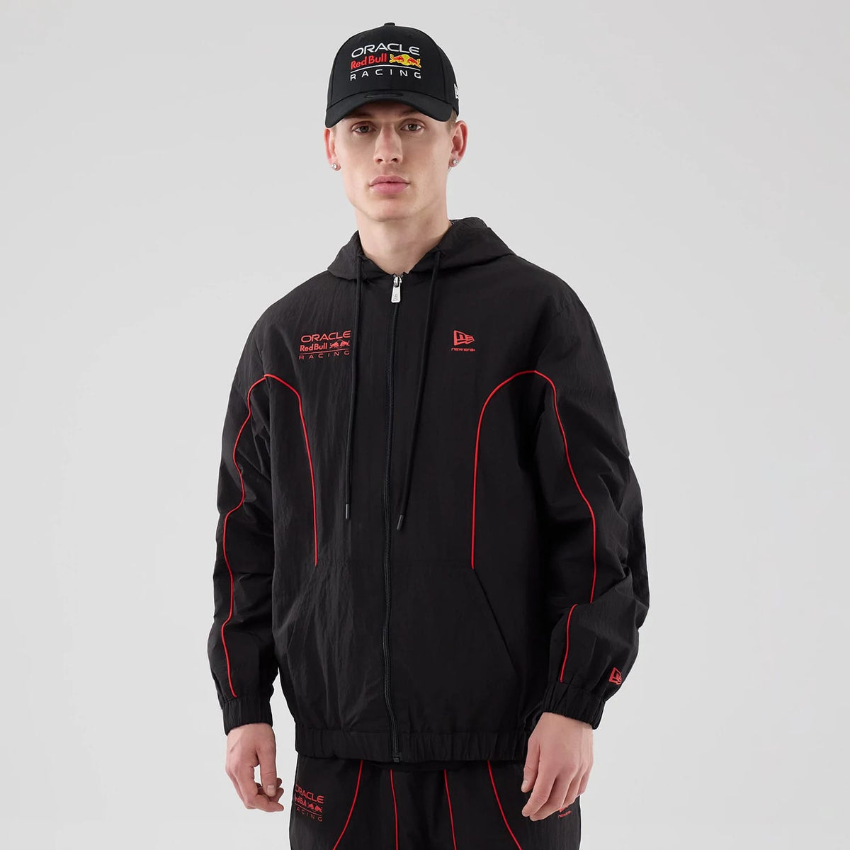 New Era Red Bull Racing Piping Black Track Jacket, front view on a model wearing a matching cap, featuring red piping along the sleeves and chest, a printed New Era logo on the upper left side and Oracle Red Bull Racing logo on the right chest.