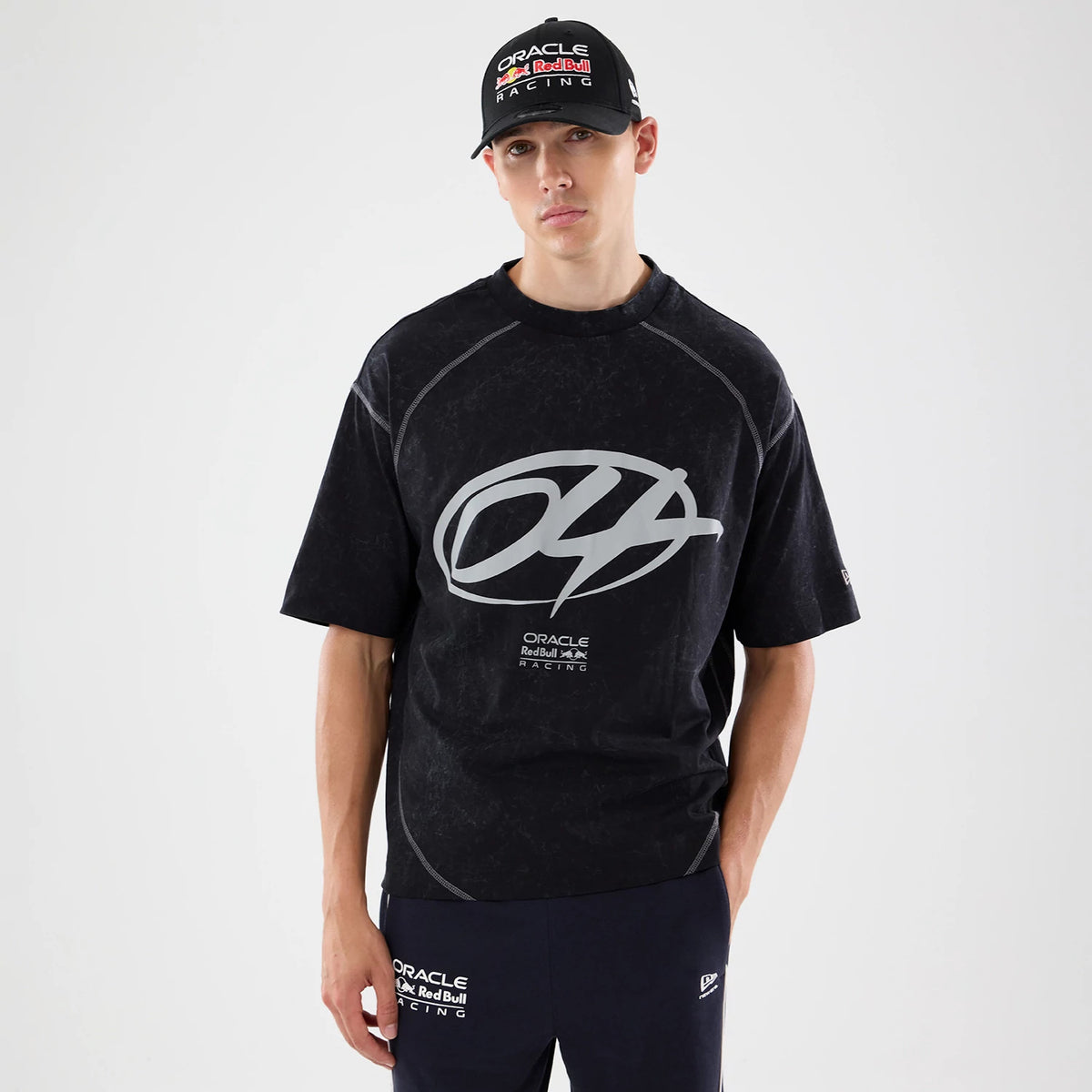 New Era Red Bull Racing Black Drop Shoulder T-Shirt, front view on a model with a matching cap, featuring a large grey graphic centred on the chest, Oracle Red Bull Racing text below, and small New Era logo embroidered on the left sleeve.