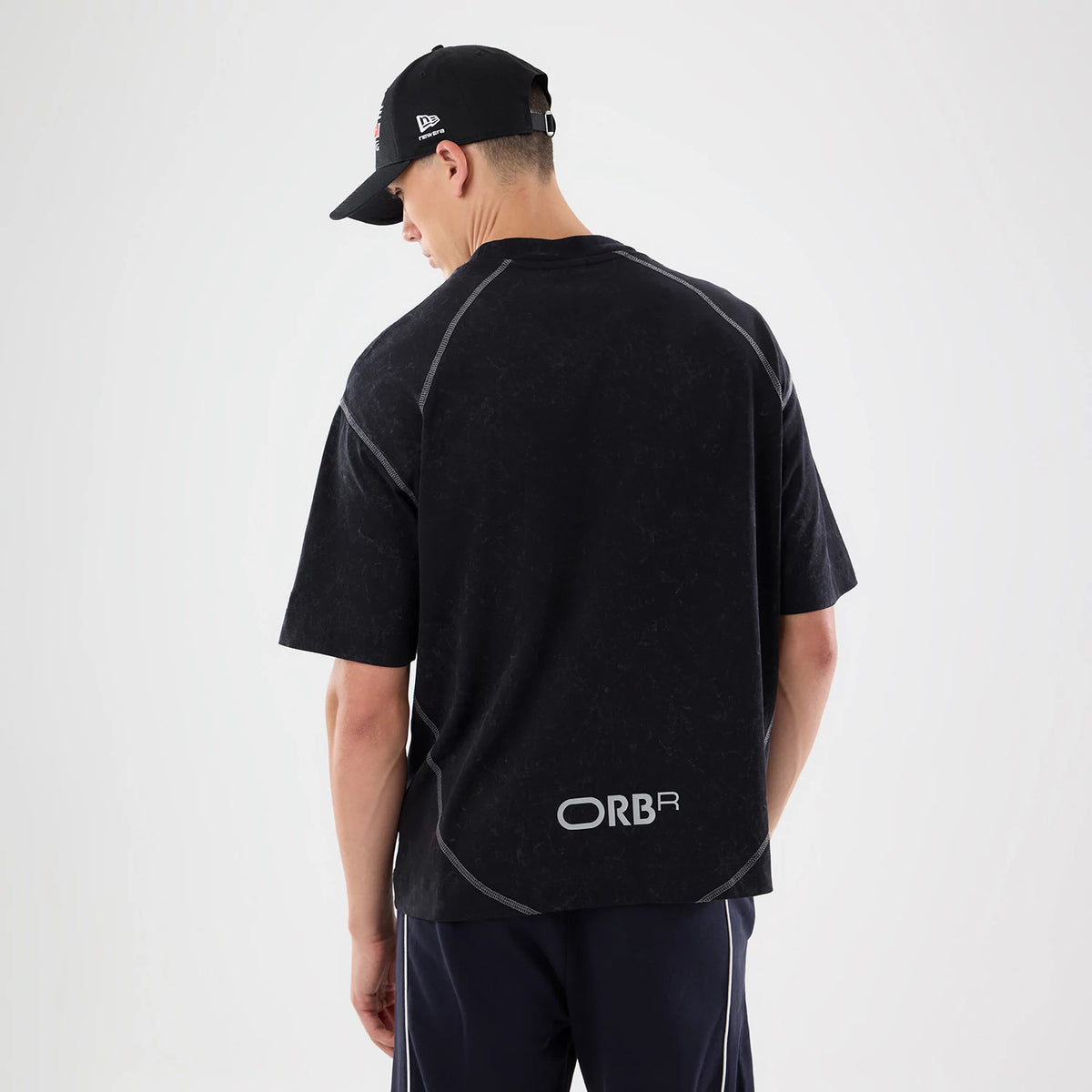New Era Red Bull Racing Black Drop Shoulder T-Shirt, back view on model displaying the ORBR printed logo near the lower hem, with seam on the drop shoulders, paired with dark trousers and a black New Era cap.