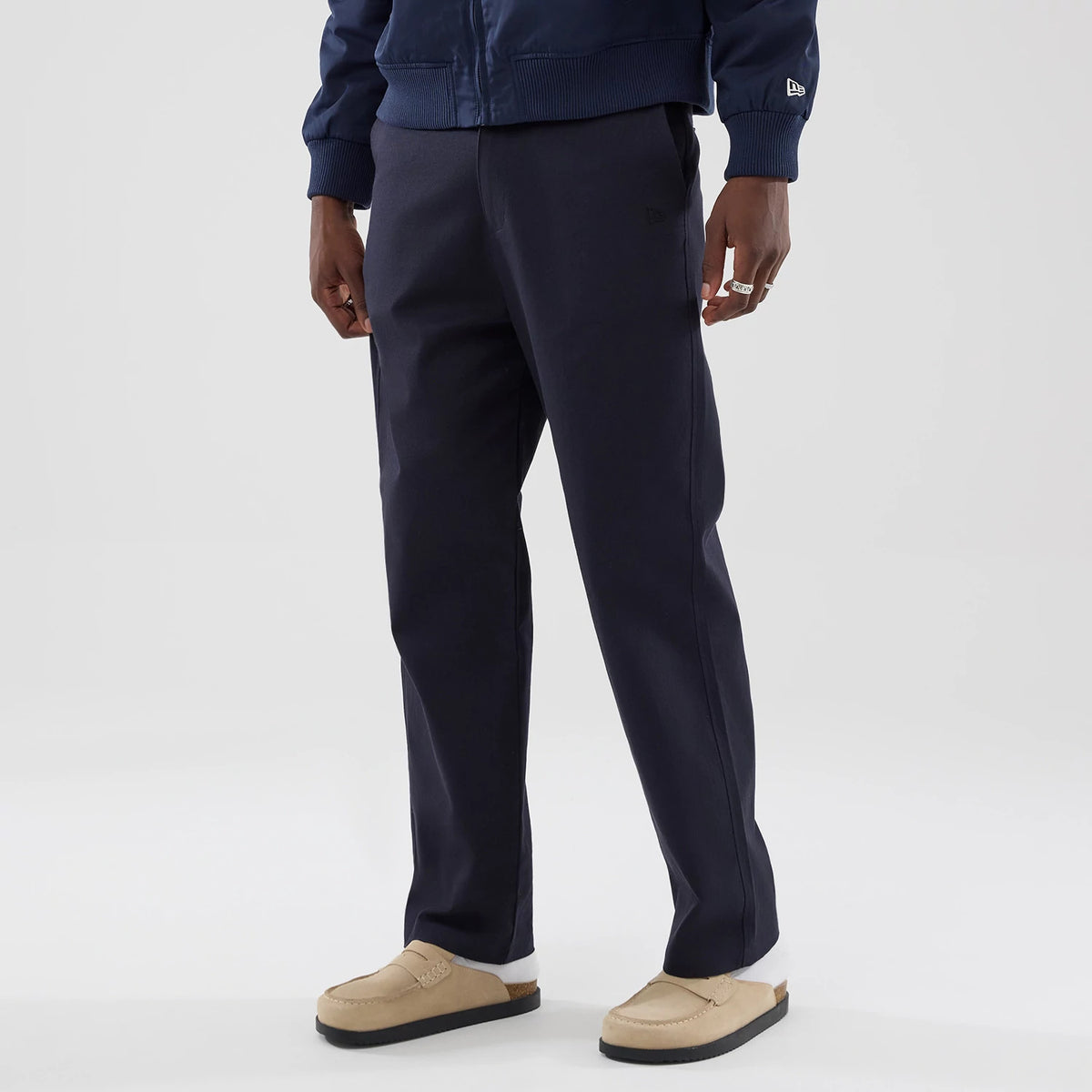 New Era Navy Chino Trousers, three-quarter view on a model wearing beige moccasins, showing the straight-leg fit, the dark navy fabric with a texture, and an embroidered New Era flag logo near the side pocket.