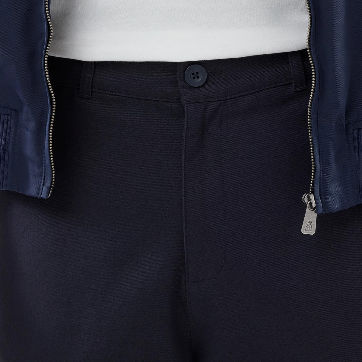 New Era Navy Chino Trousers, front view showing the dark button fastening on the waistband, a zip pull with the A logo on a jacket, and a glimpse of a white top under the open jacket.