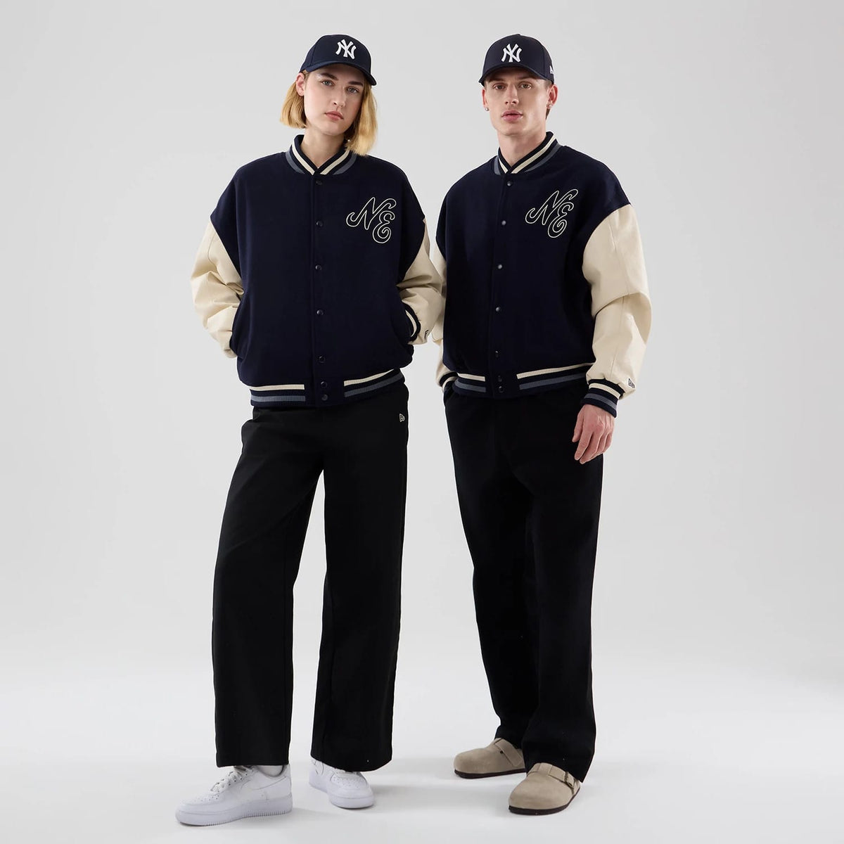 New Era Oversized Navy Varsity Jacket, front view on models wearing matching black trousers and navy caps, featuring cream-coloured sleeves, NE lettering embroidered on the chest, and striped trim details at the cuffs and waistband.