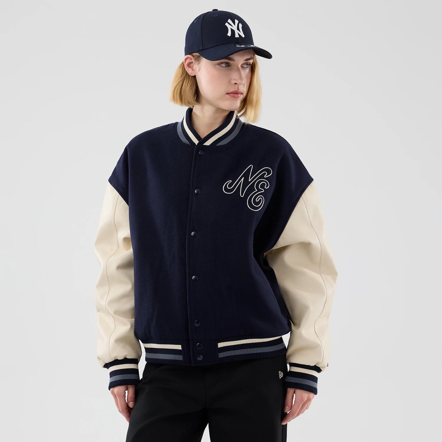 New Era Oversized Navy Varsity Jacket, front view on-model featuring a navy body with button closure, off-white sleeves, and an embroidered NE logo on the left chest, paired with dark trousers and a navy cap with a New York Yankees logo.