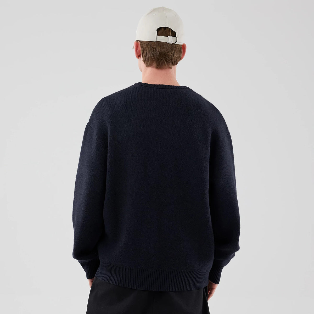 New Era Knit Script Navy Knitted Sweater, back view on a model wearing a white cap and black trousers, showing the sweater's dark navy fabric, ribbed hem and cuffs, and relaxed fit across the shoulders.