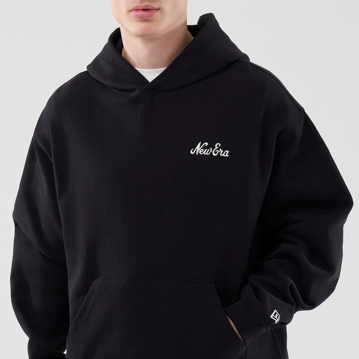 New Era Open Hem Black Pullover Hoodie, front view on a model showing a white embroidered New Era script logo on the left chest, a spacious front pocket, and a small embroidered New Era flag logo on the lower right sleeve.