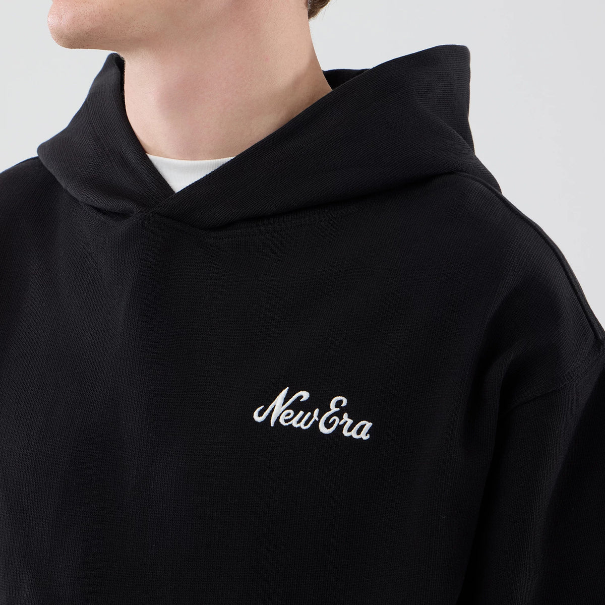 New Era Open Hem Black Pullover Hoodie, front view on-model highlighting a clean black design with an embroidered New Era script logo in white on the left chest and a spacious hood worn over a white undershirt for a casual look.