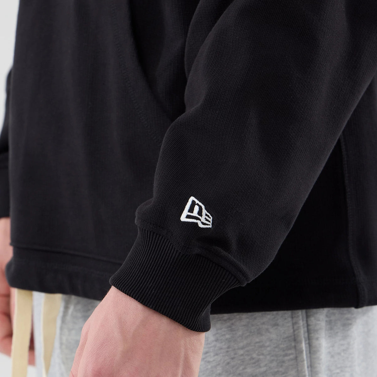 New Era Open Hem Black Pullover Hoodie, side view showing the ribbed cuff of the sleeve with an embroidered white New Era logo near the wrist, a relaxed open hem design, and part of grey trousers in the background.